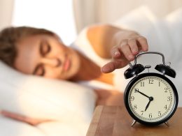 Frau mit braunen Haaren liegt im Bett und schaltet den Wecker aus