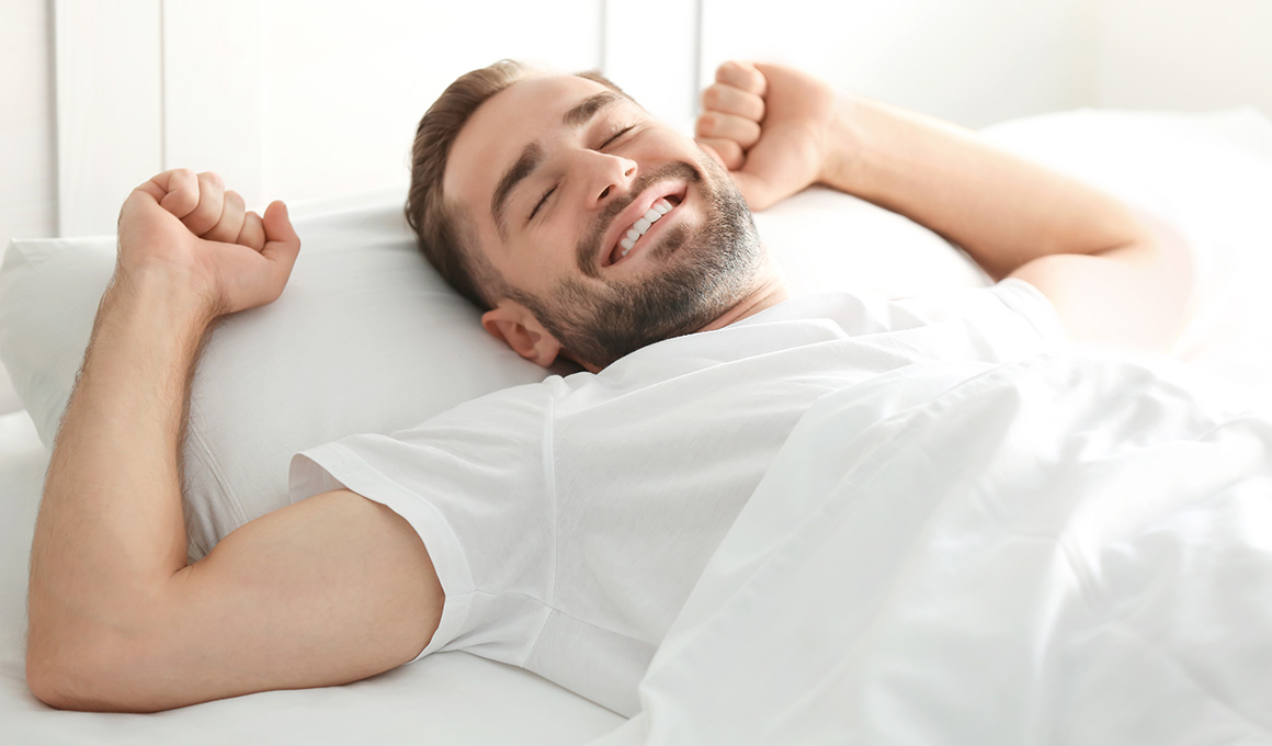 Mann im weissen T-Shirt streckt sich im Bett.