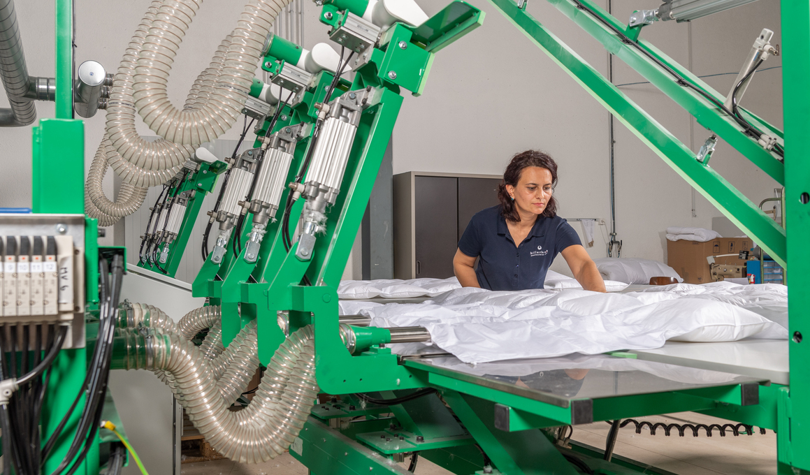 Mitarbeiterin kontrolliert die Befüllung eines Daunen-Duvets an einer industriellen Fertigungsanlage – moderne Produktion von Allergiker geeigneten Naturbettwaren.“