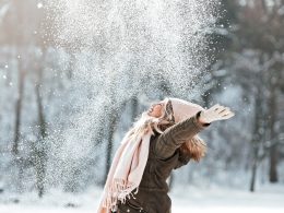 Fröhliche Frau wirbelt lachend Schnee in die Luft – Symbol für Erholung, Lebensfreude und guten Schlaf während aktiver Sportferien in winterlicher Umgebung.