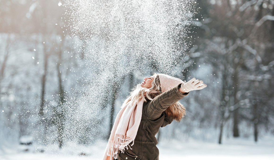 Fröhliche Frau wirbelt lachend Schnee in die Luft – Symbol für Erholung, Lebensfreude und guten Schlaf während aktiver Sportferien in winterlicher Umgebung.