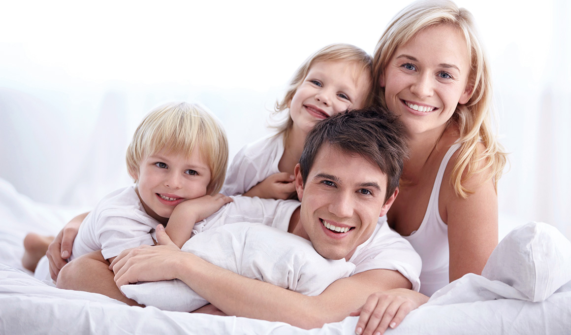 Lächelnde Familie im Bett – Symbol für erholsamen Schlaf und gesundes Leben durch passende Duvets und ergonomische Kissen.