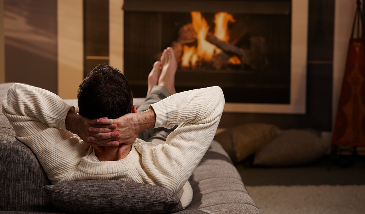 Mann liegt auf dem Sofa und wärmt seine Füsse vor dem Kamin.