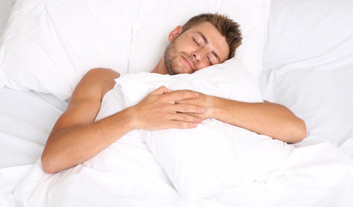 Man liegt zugedeckt in seinem Bett und hält sein Kissen umarmt.