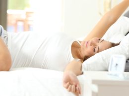 Frau mit weissem T-Shirt streckt sich im Bett um frisch und munter in den Tag zu starten.