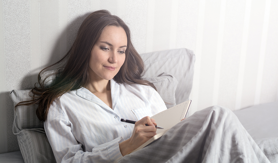 Frau mit schwarzen Haaren liegt im Bett mit ihrem Notizbuch in der Hand.