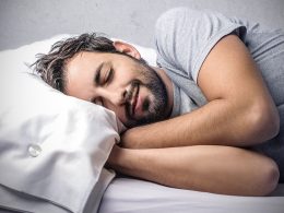 Man mit grauem T-Shirt schläft seitlich auf sein Kissen. Seine Hände sind unter dem Kissen.