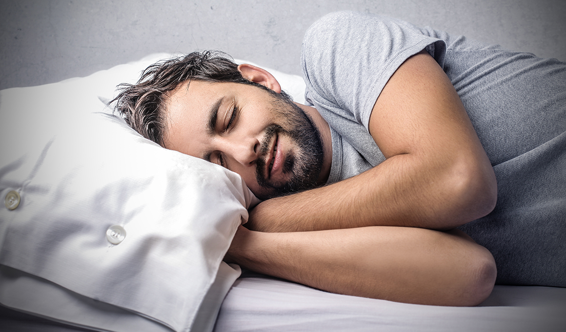 Man mit grauem T-Shirt schläft seitlich auf sein Kissen. Seine Hände sind unter dem Kissen.