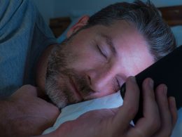 Mann schläft in seinem Bett mit seinem Handy in Hand und wird vom Handylicht beleuchtet.