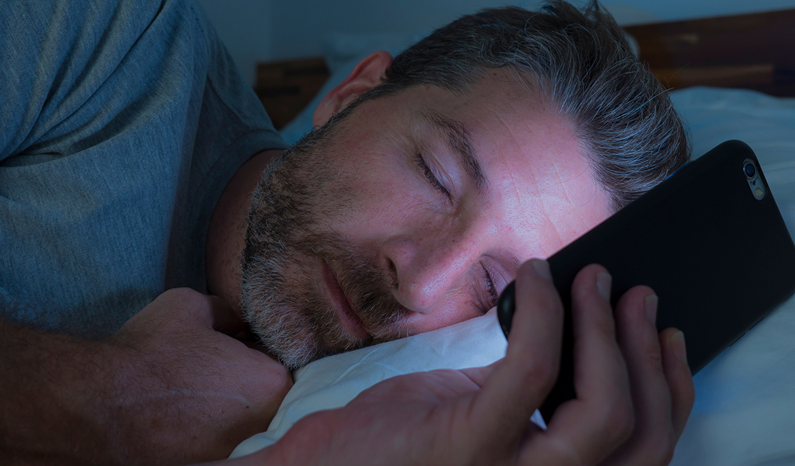 Mann schläft in seinem Bett mit seinem Handy in Hand und wird vom Handylicht beleuchtet.