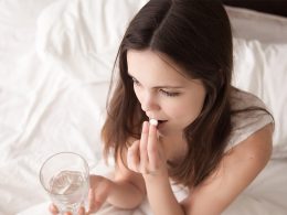 Junge Dame sitzt auf Bett und nimmt Schlafmittel ein. In der rechten Hand hält sie ein Glas Wasser.