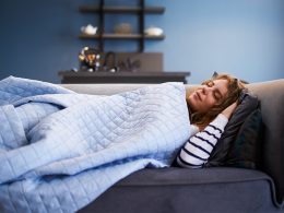 Frau mit braunen Haaren liegt zugedeckt auf ihrem grauen Sofa mit geschlossenen Augen und hält einen Mittagsschlaf