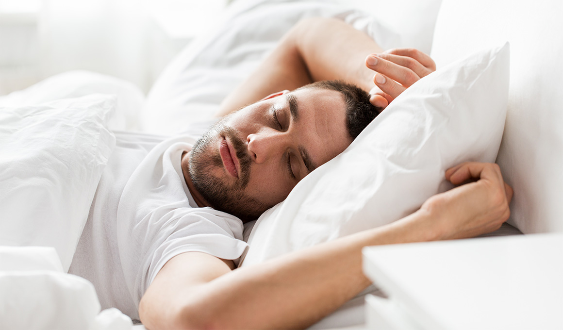 Mann mit kurzen, schwarzen Haaren in weissem T-Shirt schläft auf in seinem Bett