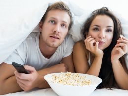 Ein Junge und ein Mädchen liegen im Bett unter der Decke und essen Popcorn und schauen Fernsehen.