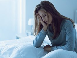 Frau hält sich die Hand übers Gesicht und sitzt müde im Bett. Ein Anzeichen dafür, dass sie Schlaf nachholen muss.