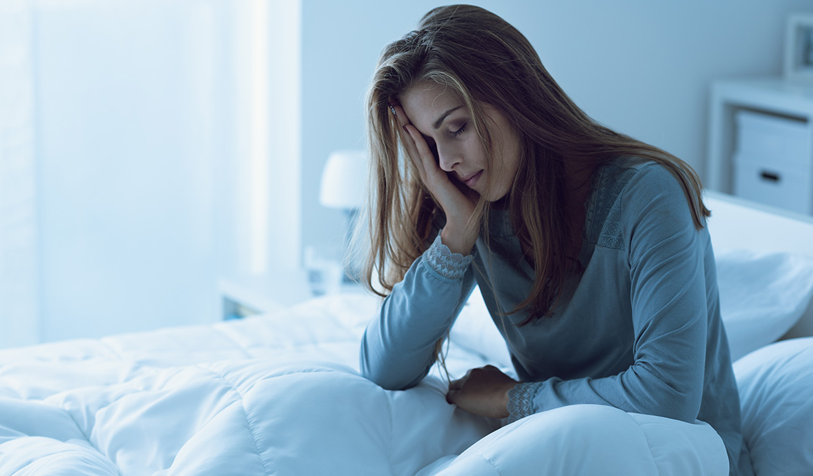 Frau hält sich die Hand übers Gesicht und sitzt müde im Bett. Ein Anzeichen dafür, dass sie Schlaf nachholen muss.