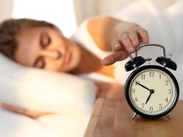 Im Vordergrund steht ein Wecker auf einem Nachttisch, Im Hintergrund liegt eine Frau im Bett und streckt eine Hand nach dem Wecker, um ihn abzustellen.