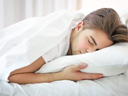 Junger Mann schläft bäuchlings im Bett. Er ist mit einer dünnen weissen Decke zugedeckt, es ist nur der Kopf und ein Arm sichtbar.