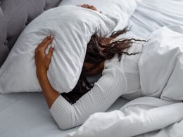 Frau liegt auf dem Bauch im Bett und deckt ihren Kopf mit dem Kissen zu. Sie hält das Kissen seitwärts mit beiden Händen.