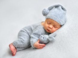 Neugeborenes Baby im grauen Wollanzug und mit grauer Zipfelmütze schläft mit einem Stoffhasen im Arm.