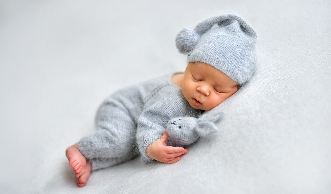 Neugeborenes Baby im grauen Wollanzug und mit grauer Zipfelmütze schläft mit einem Stoffhasen im Arm.