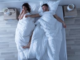 Junges Paar schläft im Bett. Der Mann liegt mit breit ausgestreckten Armen und Beinen auf dem Rücken und braucht den meisten Platz. Die Frau liegt ganz an den Rand gedrängt auf der Seite und blickt verärgert.