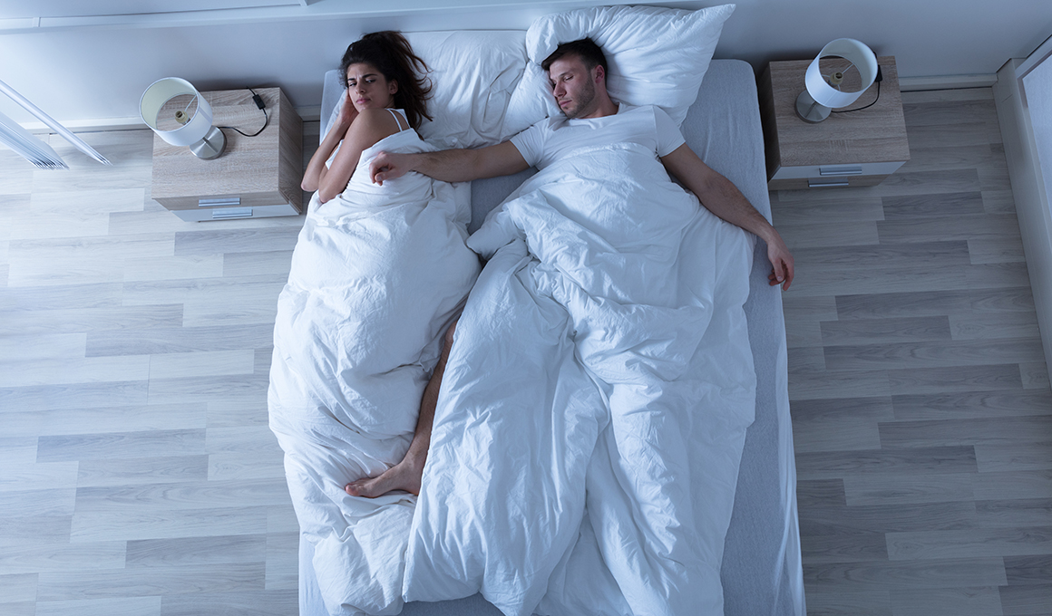 Junges Paar schläft im Bett. Der Mann liegt mit breit ausgestreckten Armen und Beinen auf dem Rücken und braucht den meisten Platz. Die Frau liegt ganz an den Rand gedrängt auf der Seite und blickt verärgert.
